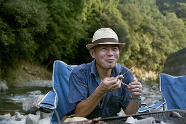 Ein Mann sitzt auf einem Camping-Stuhl vor Bäumen und einem Fluss.
