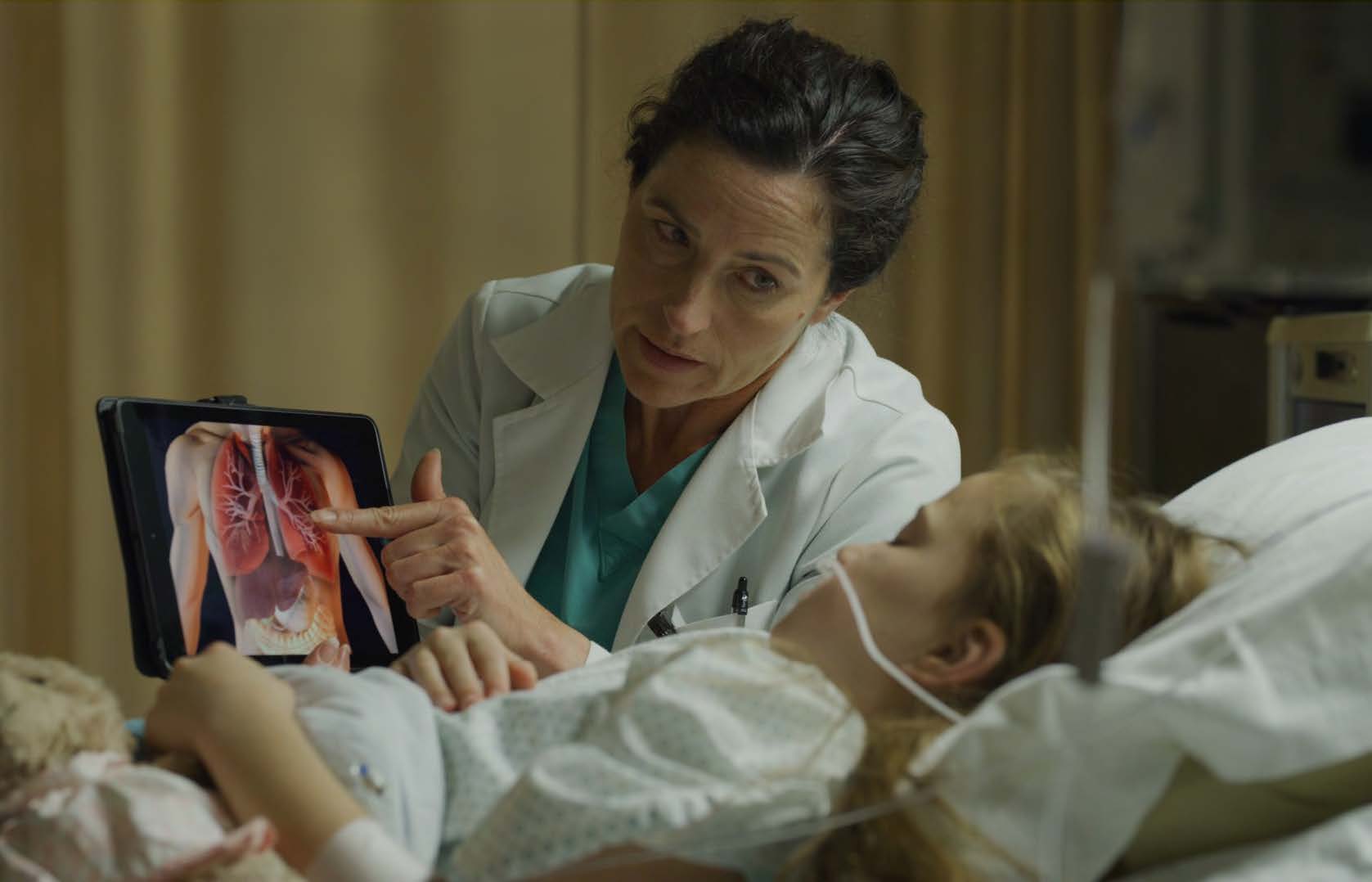 Uma menina loira está acamada e entubada. Ela olha com atenção para o ecrã de um tablet onde é visível um tronco humano e dentro dele os pulmões, em destaque. O tablet é segurado por uma senhora morena envergando bata branca.