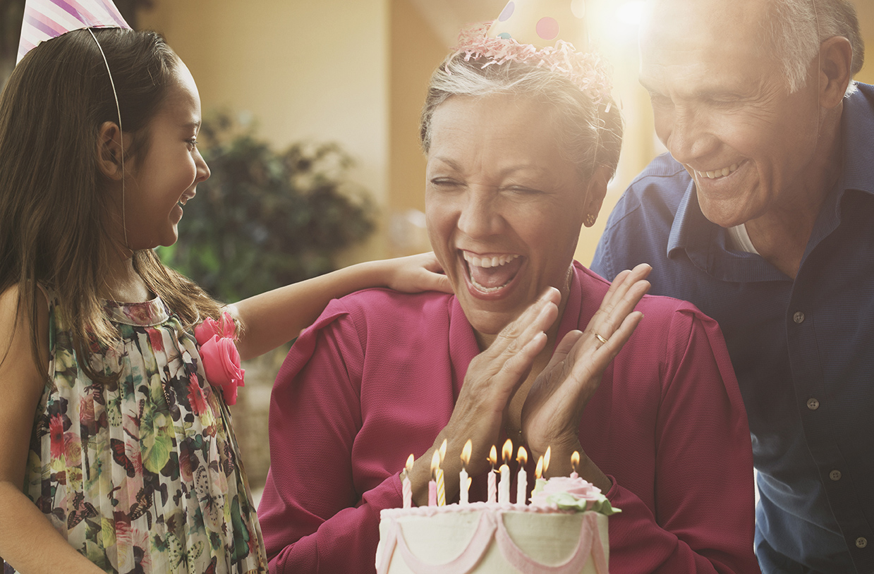 Uma mulher de meia idade muito sorridente bate palmas para o bolo de aniversário à sua frente. Está acompanhada, em cada lado, por um senhor de uma menina, que a olham também sorridentes. Todos utilizam chapéus de papel bicudos na cabeça.