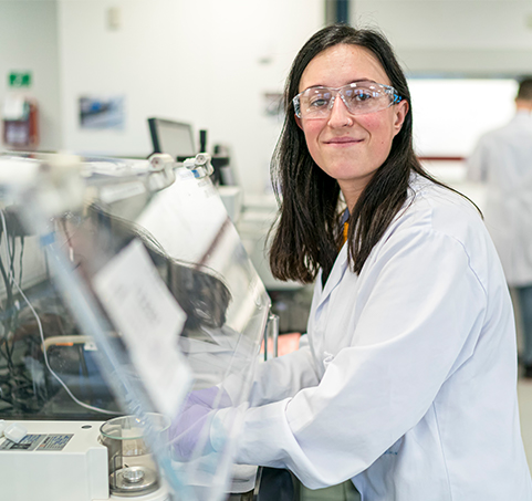 Mulher cientista, vestida de bata branca e usando óculos de proteção, sorri enquanto manipula equipamentos técnicos num laboratório.