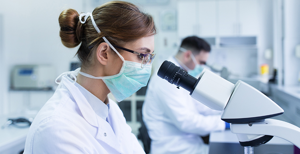 Mulher cientista, vestida com bata branca e a usar máscara cirúrgica e óculos, olha pela lente de um microscópio numa mesa de laboratório onde está sentado outro profissional.